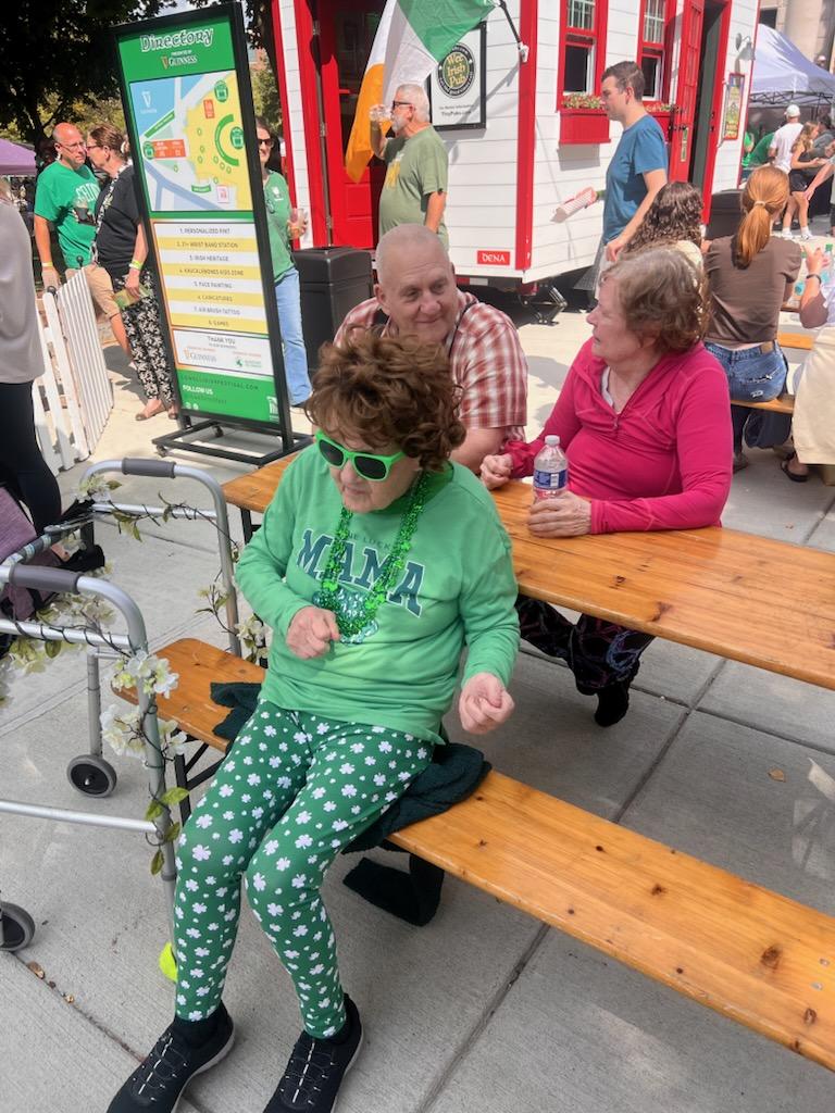 A woman in green sits on a bench outdoors while people gather around