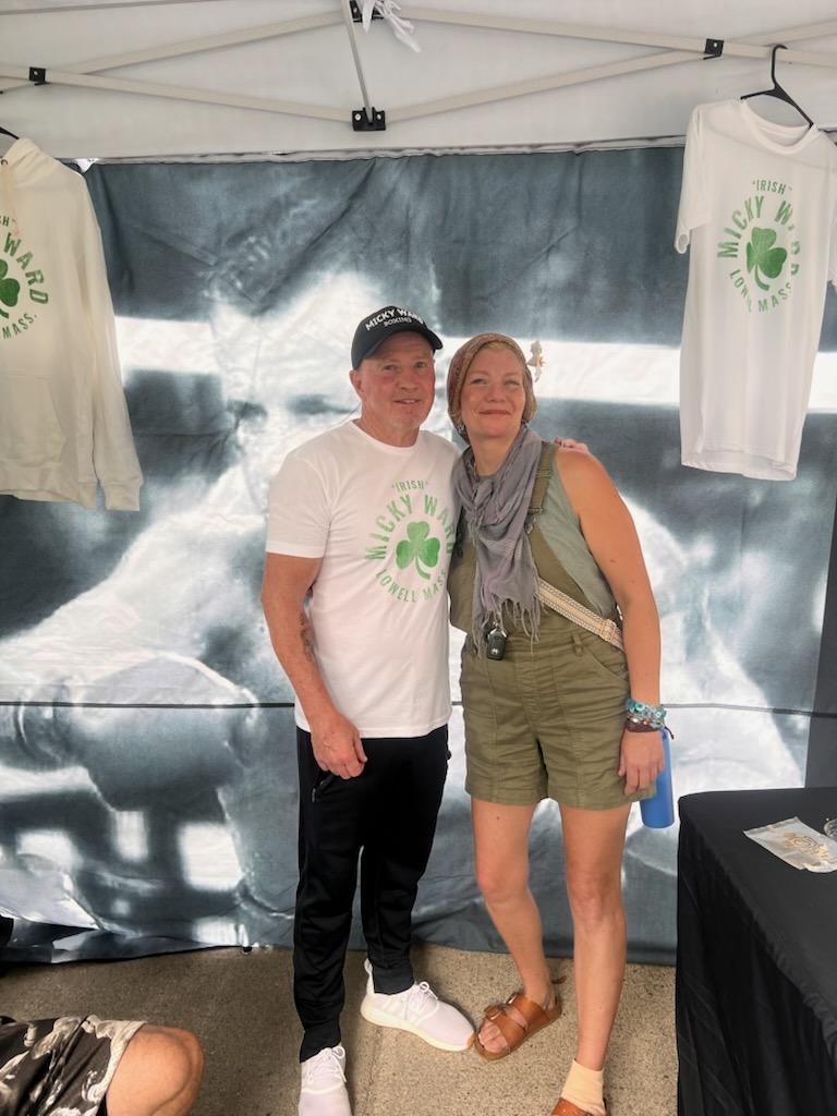 Two people pose together under a tent with a shirt display behind them.